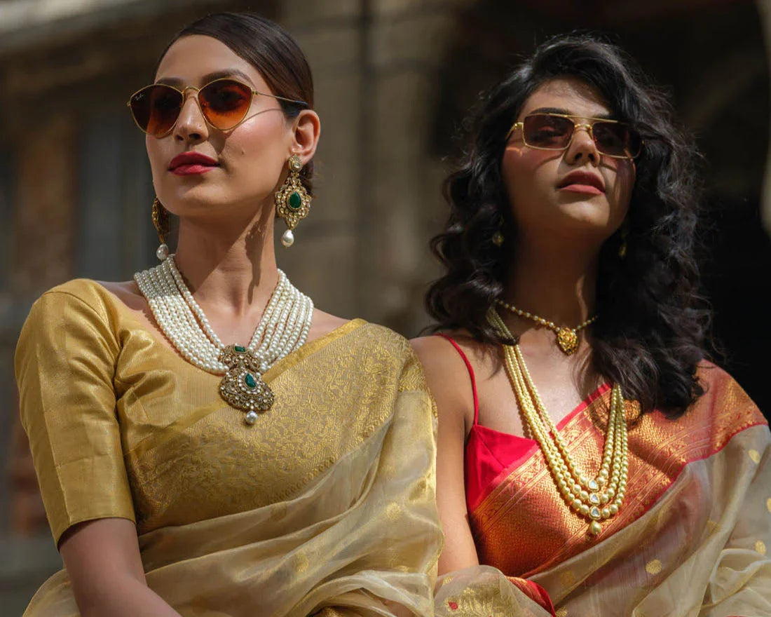 Two women wearing premium handloom sarees and gold pearl jewelry, styled with sunglasses in natural light