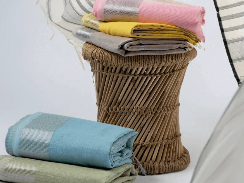 Stack of colorful handloom sarees in pastel pink, yellow, grey, blue, and green on a wicker stool, white background