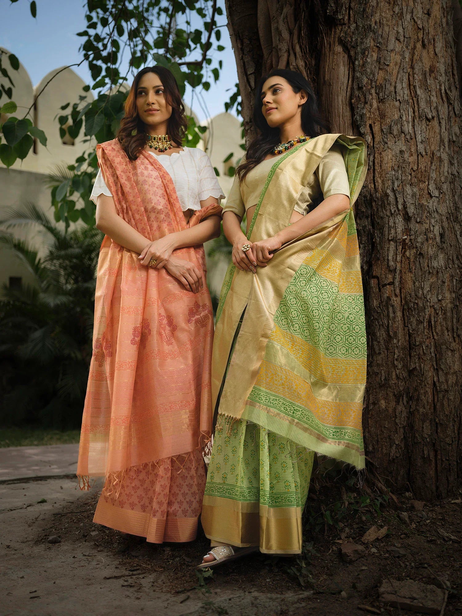 Two women wearing elegant handloom sarees, one in peach and white, the other in green and gold, standing by a tree outdoors