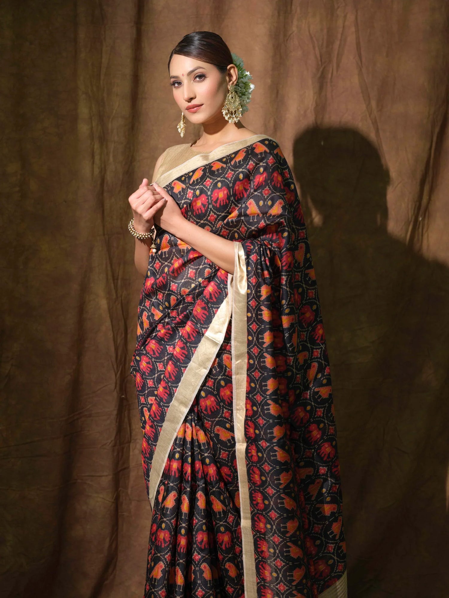 Woman wearing a premium handloom saree with red and orange elephant motifs on black, gold border, styled with statement earrings and flower hair accessories